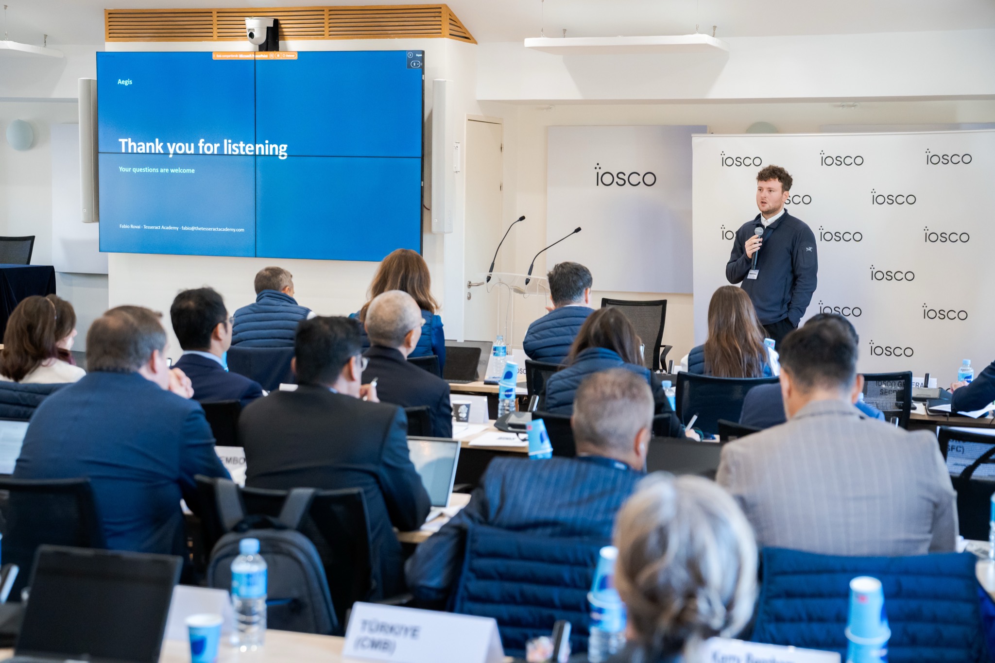 Fabio Rovai of Tesseract Academy presents Aegis to delegates at the IOSCO TechSprint Demo Day in Madrid, standing in front of IOSCO banners beside the closing slide